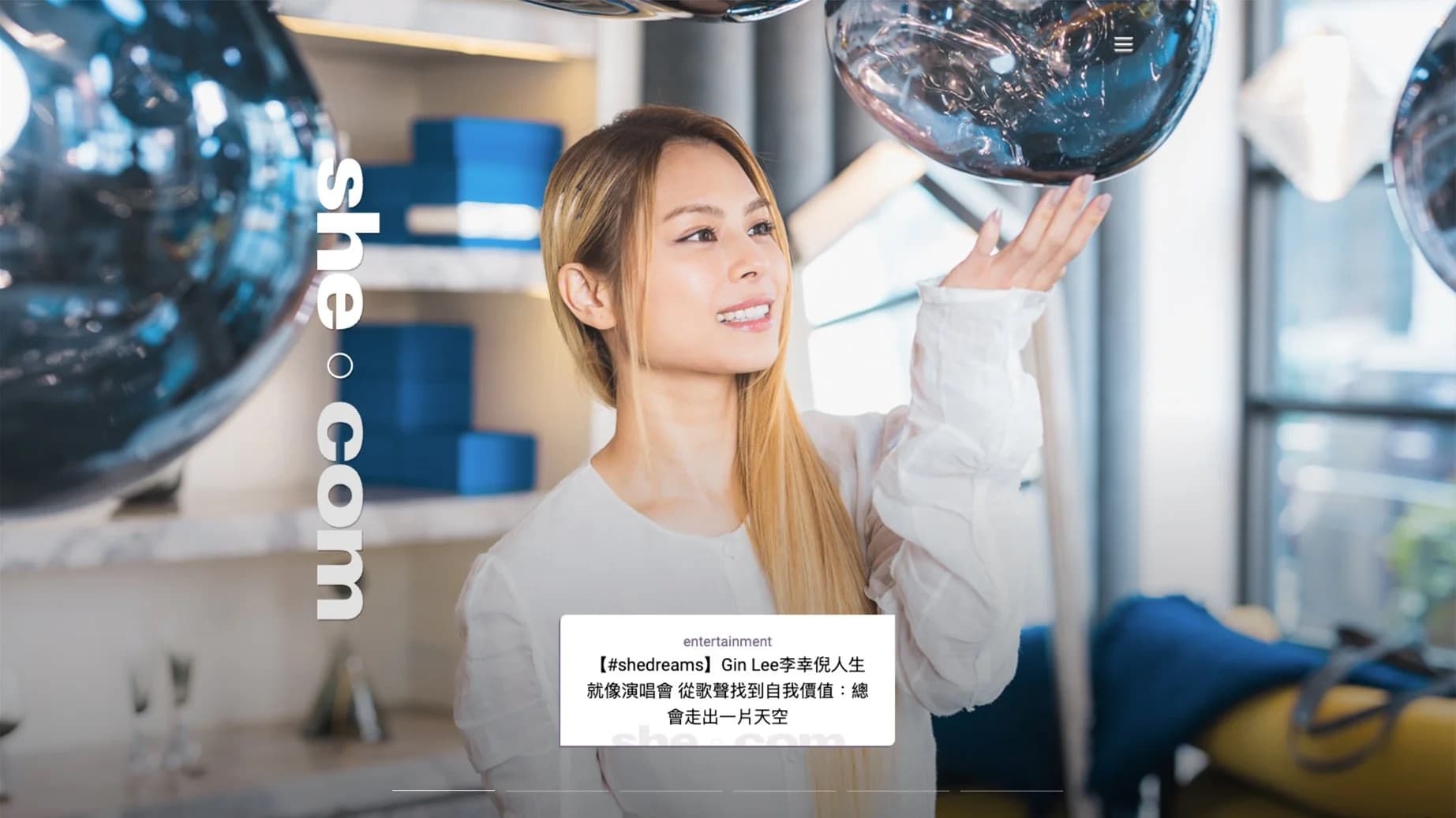 A woman with long blonde hair adjusting or examining large black balloons in a bright, modern room with shelves and glass windows.